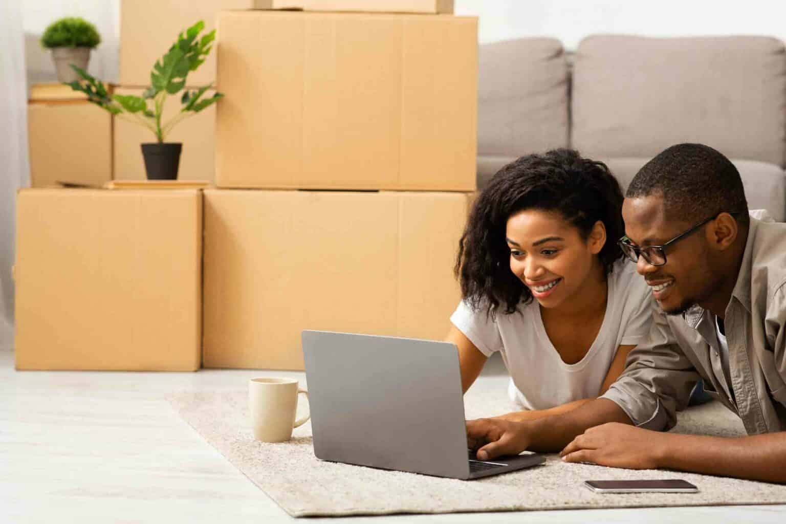 a couple sitting on the floor looking at insurance agency options on their laptop with moving boxes in the background