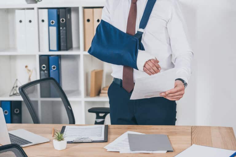 cropped view of worker with broken arm in bandage standing and holding workers' compensation form over table in office,