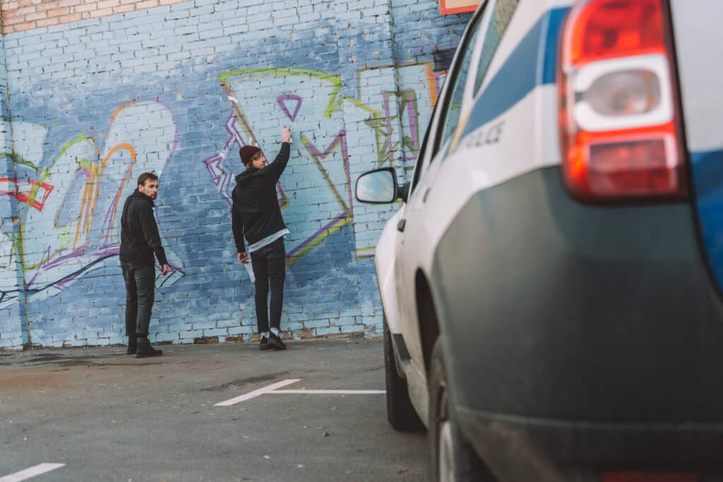 back view of vandals painting graffiti on wall with a police car in the foreground