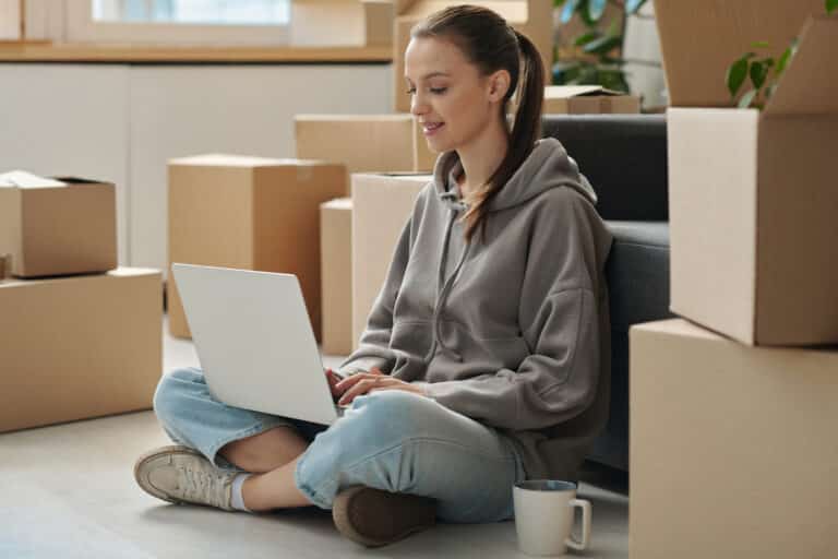 Young woman using laptop to rent a new apartment while sitting on the floor among packed boxes during relocation