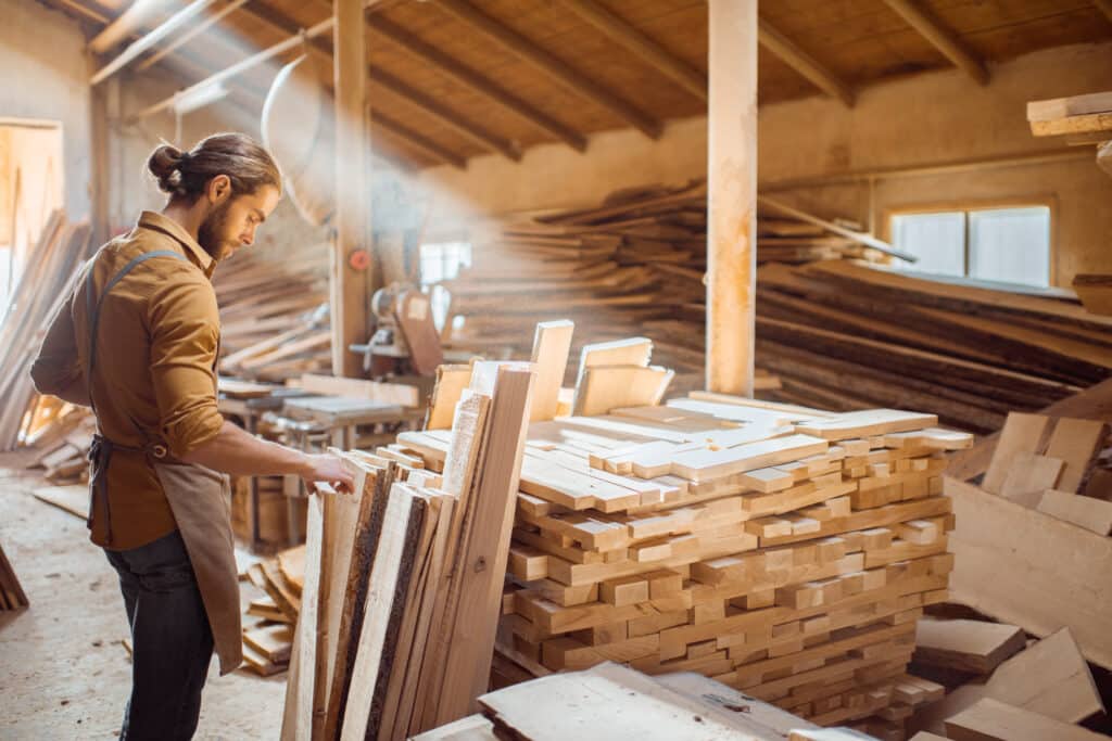 A skilled contractor examines wooden planks in a well-lit woodworking shop filled with stacks of lumber. This image highlights the importance of carpenter insurance for professionals working in construction and woodworking environments, ensuring protection against potential risks and liabilities.