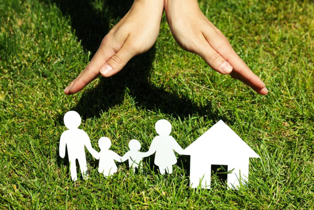Protecting family and home on green grass with hands sheltering paper cutouts.
