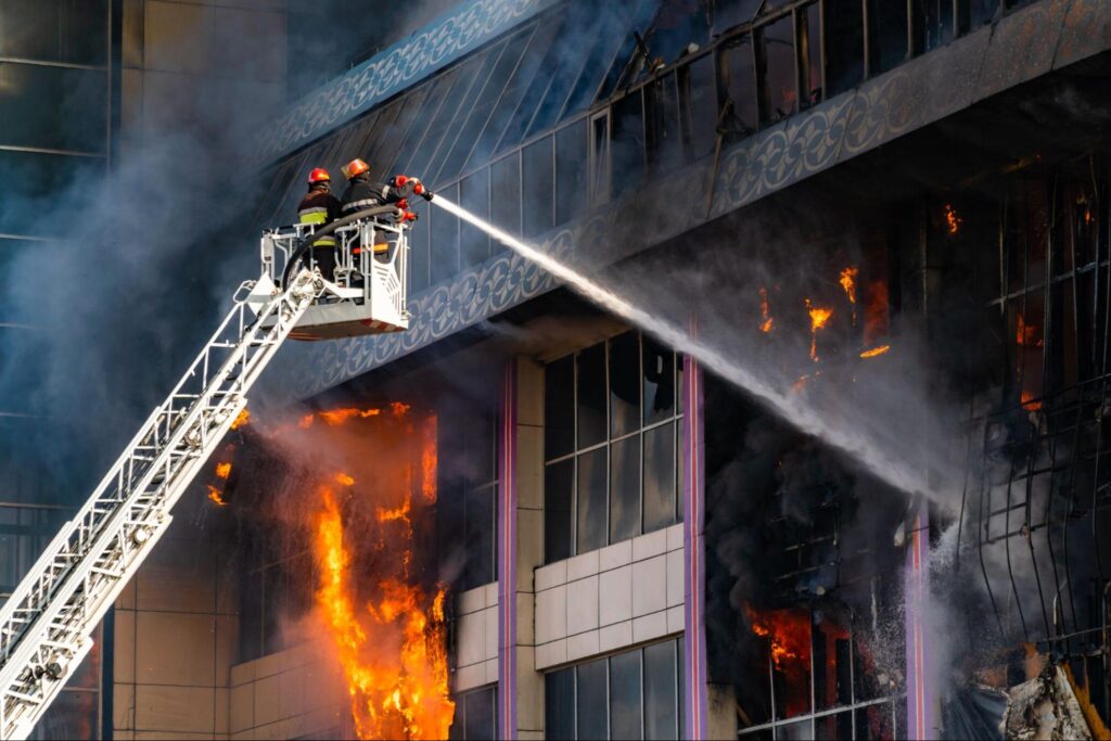 Firefighters battling a massive building fire with a ladder and hose.