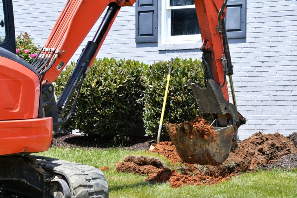 Excavator working on landscaping in a front yard, digging soil and preparing the area.