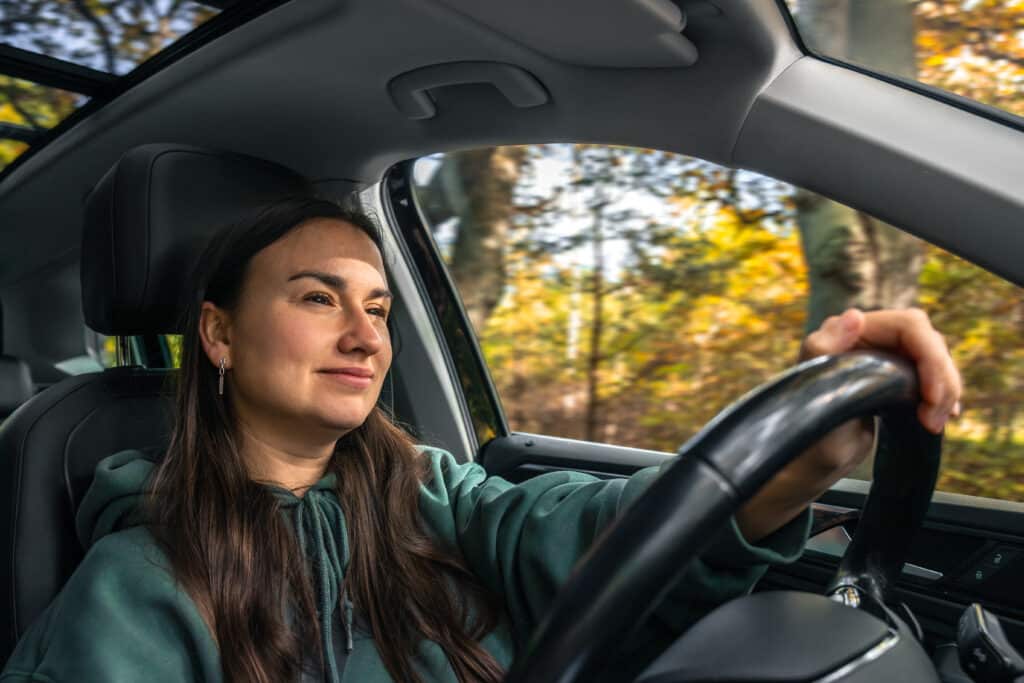 Smiling woman driving a car through a sunny forest, enjoying natures beauty.